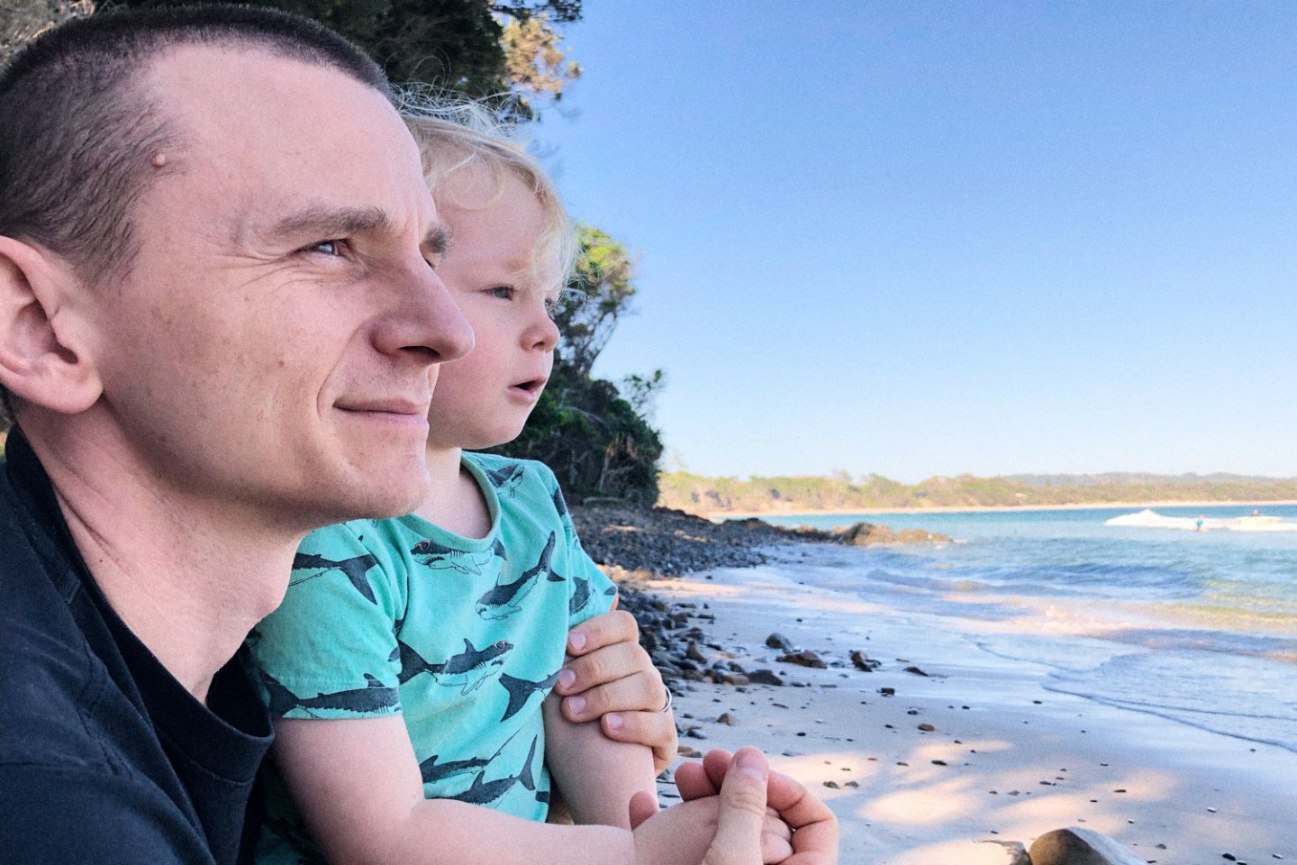 Family photos at the beach in Sydney A father wraps his arms around his young child at the beach as they both gaze out over the ocean.